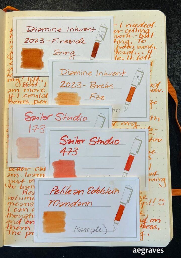 Five ink sample cards displaying orange inks atop a cream colored page of writing.  Inks include Diamine Fireside Snug, Diamine Bucks Fizz, Sailor Studio 173 & 473, and Pelikan Edelstein Mandarin.